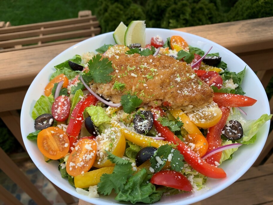 A plate with a romaine salad topped with bell peppers, tomatoes, olive, onion, cheese and a tortilla crusted fish filet. 