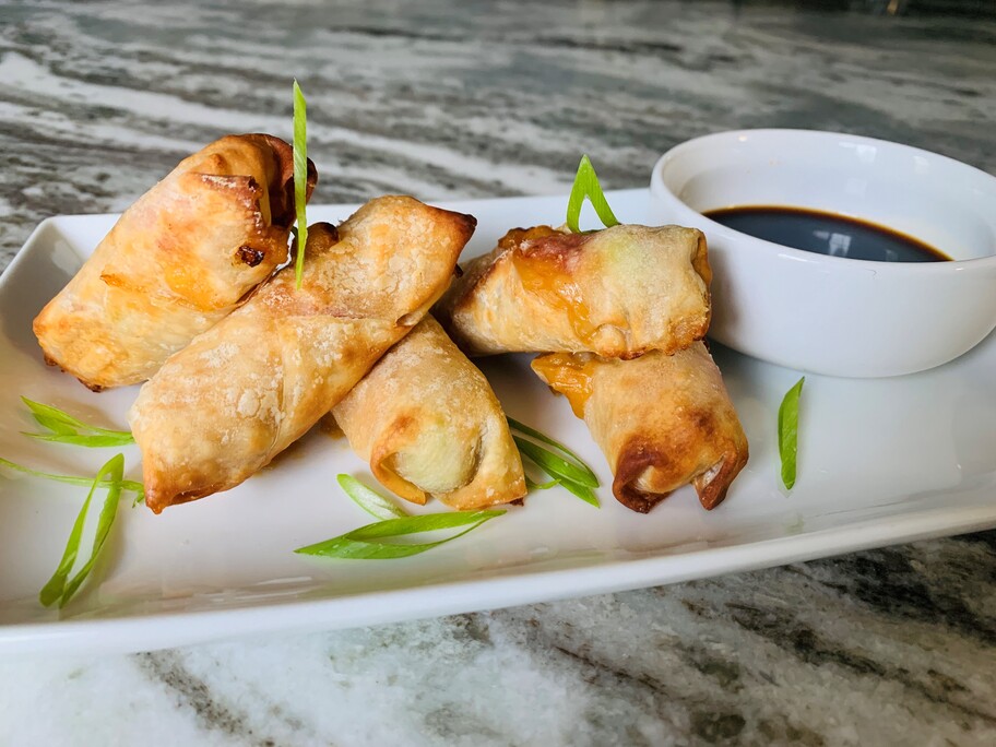 Four orange chicken spring rolls on a plate with green onion garnish and a bowl of dipping sauce.