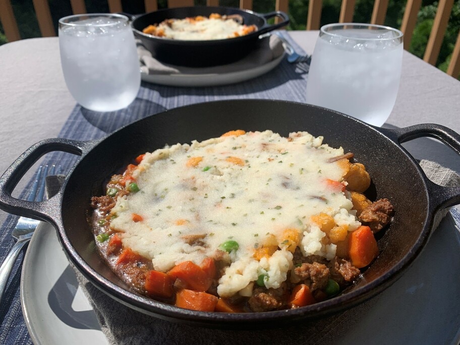 A patio table set with glasses of water, a tablecloth, napkins and two cast iron dishes full of shepherds pie.