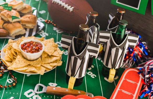 Una mesa servida para ver el partido con minihamburguesas, botellas de cerveza con cubiertas de camiseta de referí y chips y salsa sobre una mesa con una pelota de fútbol.