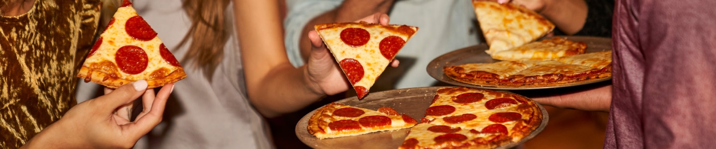 Pizza de pepperoni y queso en bandejas, y al fondo personas tomando rebanadas.