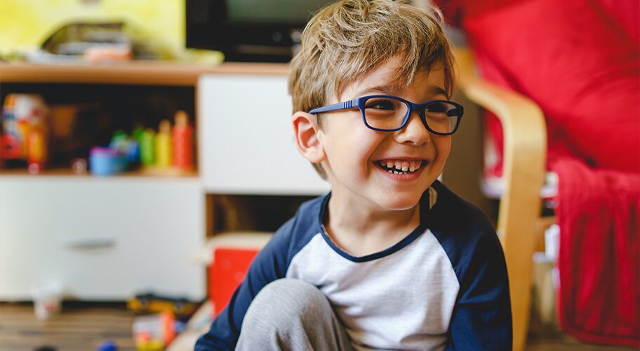 Niño con gafas sonriendo en el suelo de un salón con juguetes.