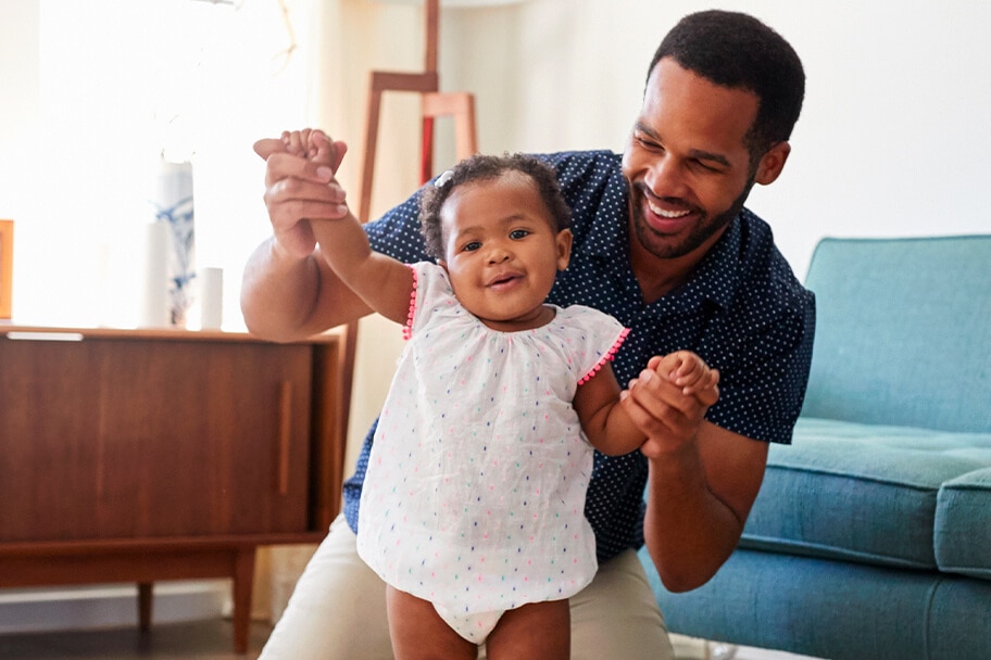 Child smiling with their father who is aiding them in walking upright in a living room.